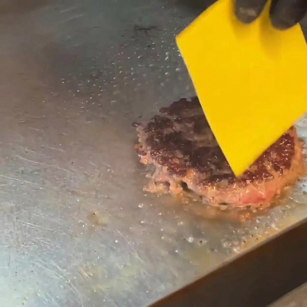 Una loncha de queso amarillo derritiéndose sobre una hamburguesa dorada usando un soplete de cocina.