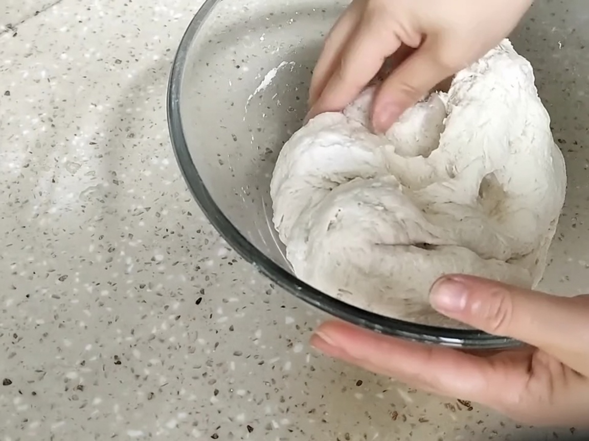 Manos presionando y amasando una bola de masa rugosa dentro de un tazón de vidrio.