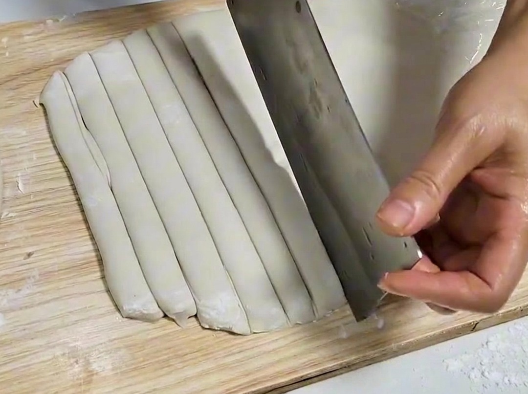 Hoja de masa cortada en tiras uniformes con una cuchilla de metal sobre una tabla de madera.