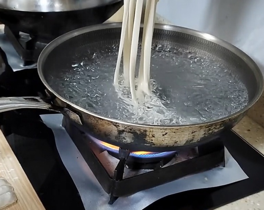 Mano estirando una tira de masa en un fideo largo antes de soltarlo en agua hirviendo.