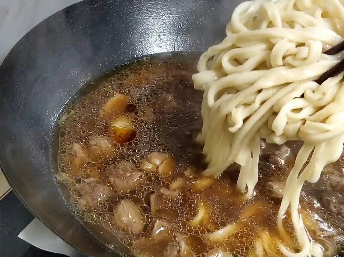 Fideos gruesos siendo levantados con palillos desde una olla de agua y colocados en un wok con la base de sopa.