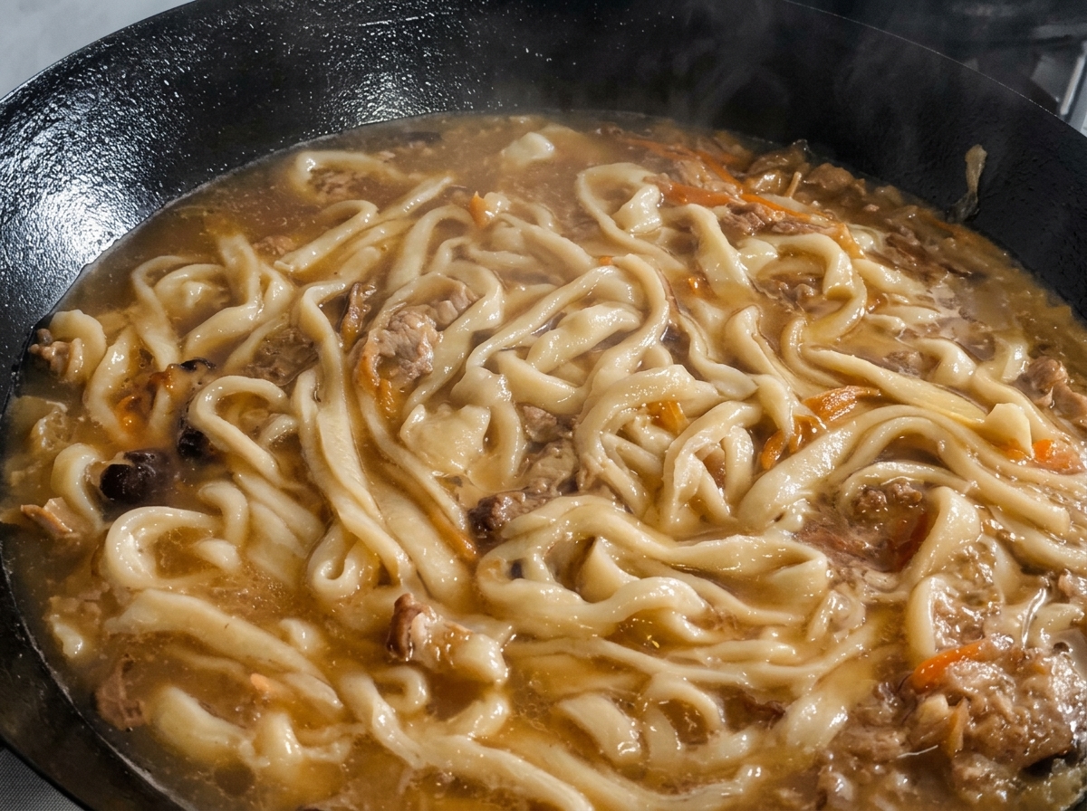 Hojas de perilla picadas siendo esparcidas sobre los fideos y la sopa hirviendo en un wok.