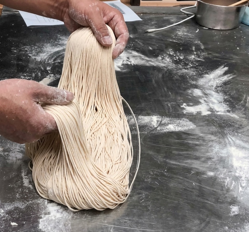 Hebras de fideos recién estirados, largas y delgadas, sostenidas en el aire.