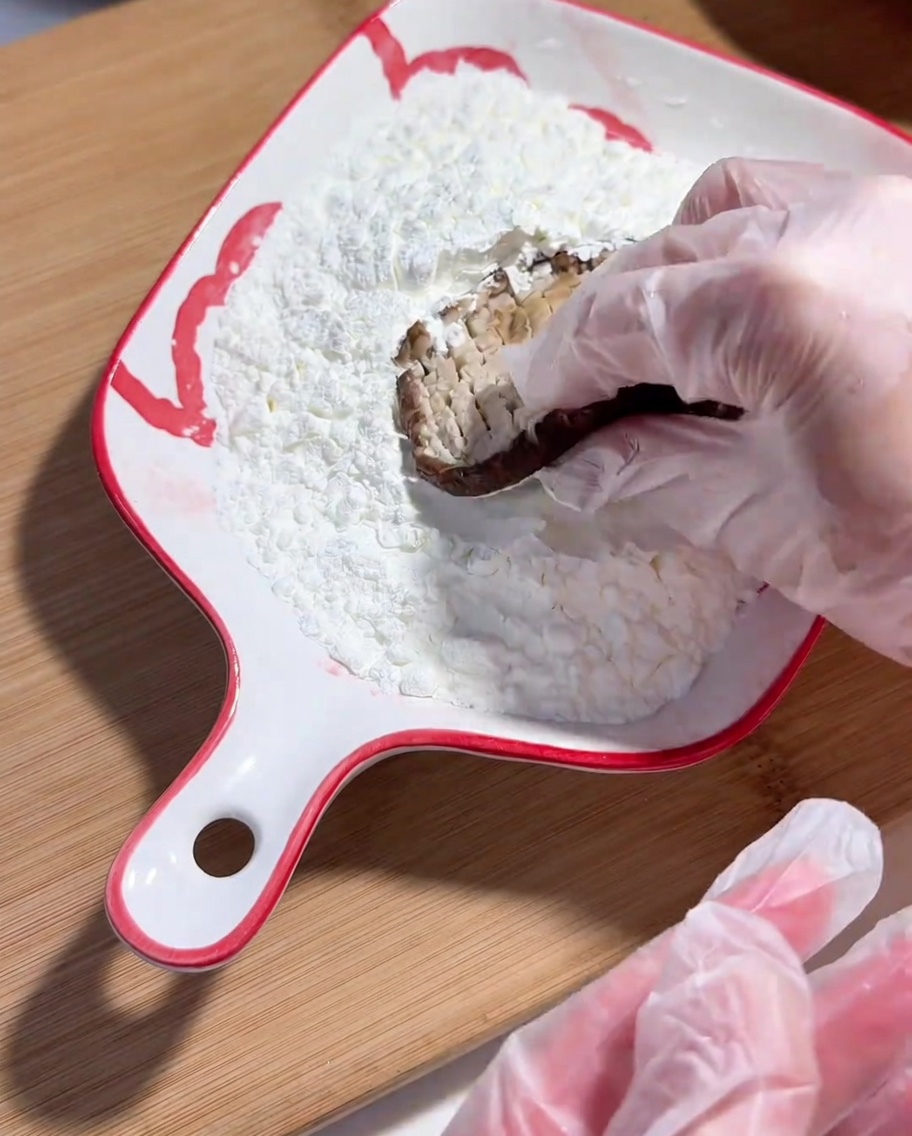 Una mano con guante de plástico sumergiendo el lado marcado de un champiñón shiitake en un plato con almidón de maíz.