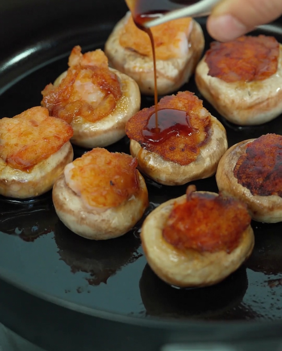 Vertiendo una mezcla de salsa de soja y jugo de champiñón sobre los champiñones rellenos en la sartén.