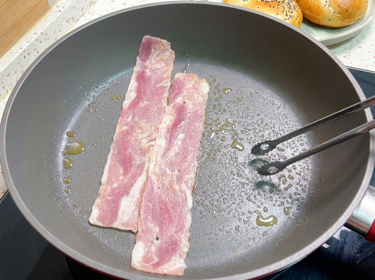Tiras de beicon friéndose en una sartén caliente, con unas pinzas volteando una de las rodajas ligeramente doradas.