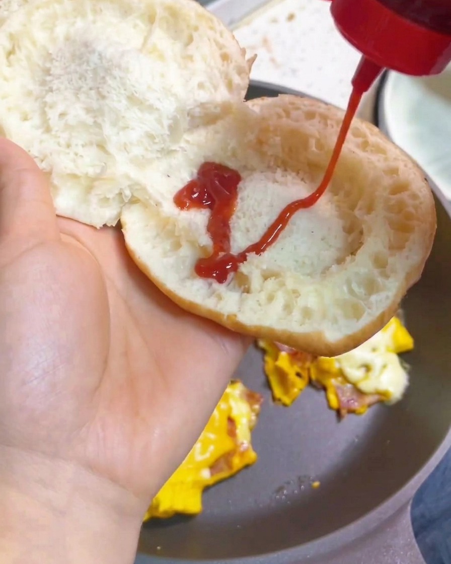 Una mano aplicando un patrón de zigzag de kétchup rojo brillante sobre el interior de un pan de hamburguesa tostado.