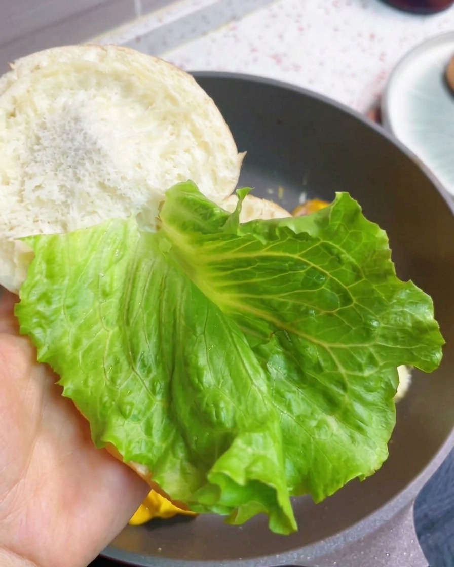 Una hamburguesa de desayuno montándose con una hoja de lechuga fresca y la tortita de beicon, huevo y queso dentro del pan tostado.