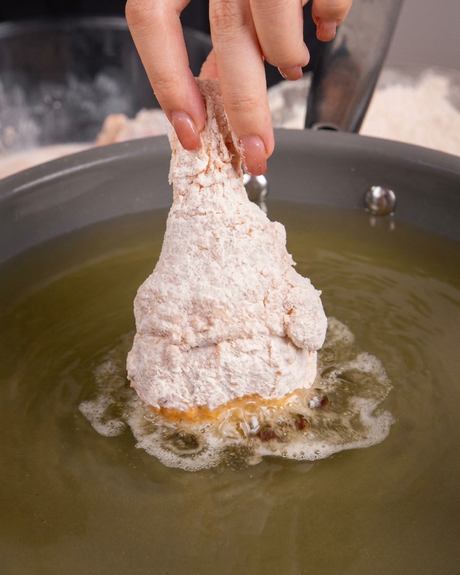 Muslo de pollo crudo y empanizado sumergiéndose en aceite caliente en una sartén.
