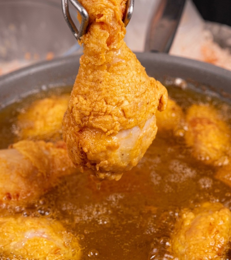 Un solo muslo de pollo frito dorado sostenido por pinzas de metal sobre una sartén de aceite caliente burbujeante.