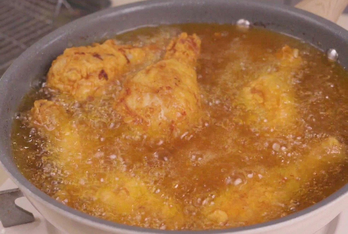 Piezas de pollo frito siendo sumergidas en una sartén con aceite caliente burbujeante para la etapa final de fritura.