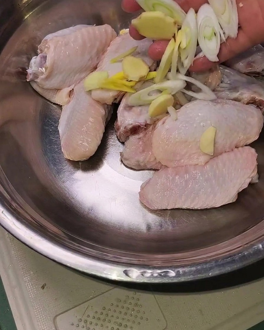 Alitas de pollo crudas en un bol de metal cubiertas con jengibre en rodajas, ajo y cebollino listas para sazonar.