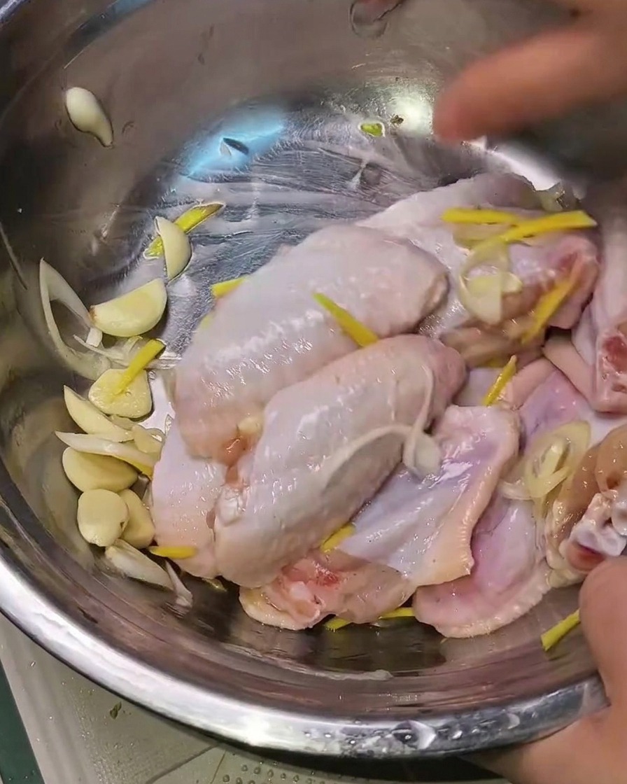 Manos mezclando las alitas de pollo marinadas con aromáticos en un bol de metal.