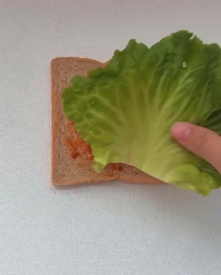 Dos hojas grandes de lechuga verde siendo colocadas sobre una rebanada de pan ya cubierta con salsa roja.