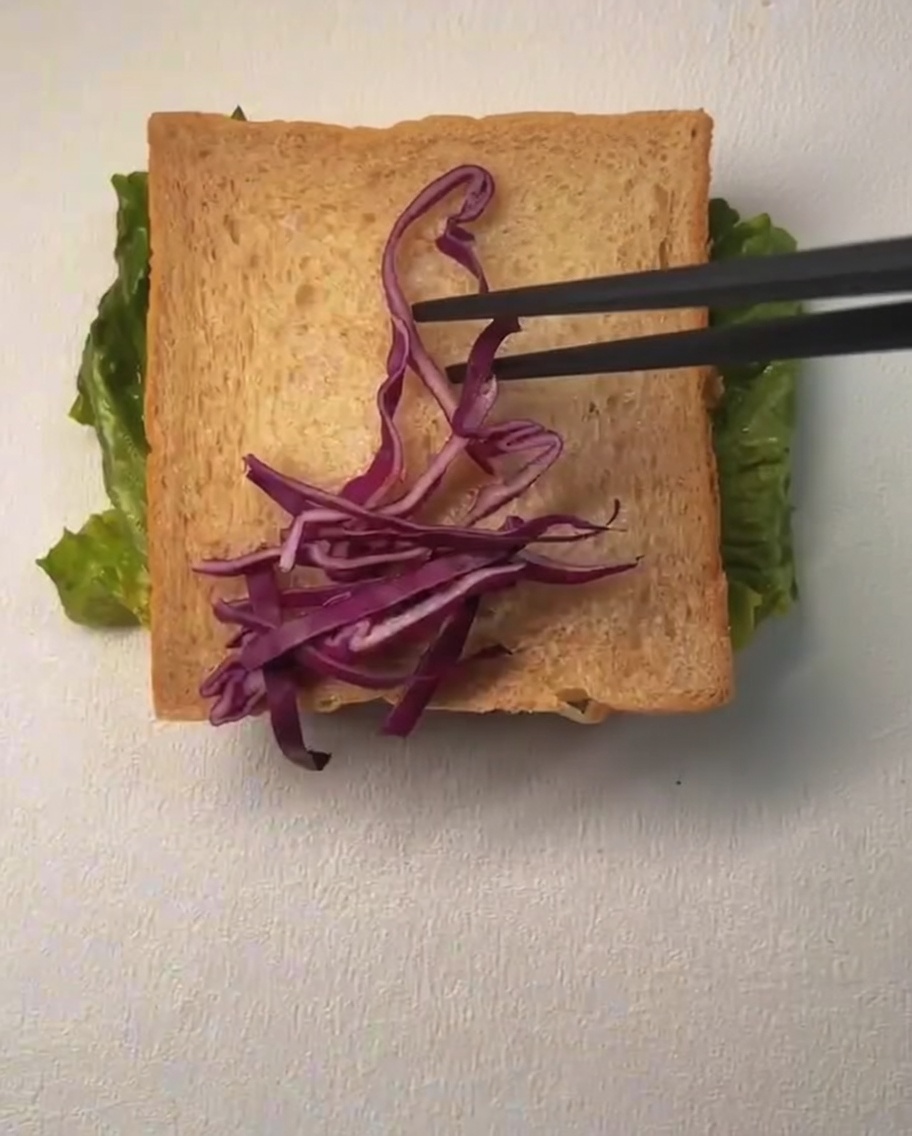 Una rebanada de pan cubierta con col morada rallada y granos de maíz, con mayonesa siendo rociada por encima.
