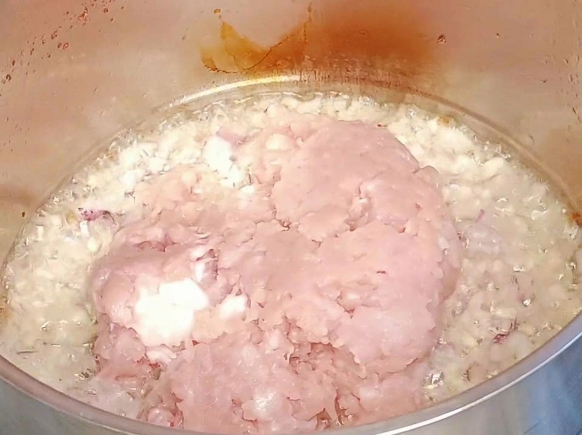 Carne molida cruda siendo añadida sobre la cebolla y el ajo sofritos en una olla de metal.