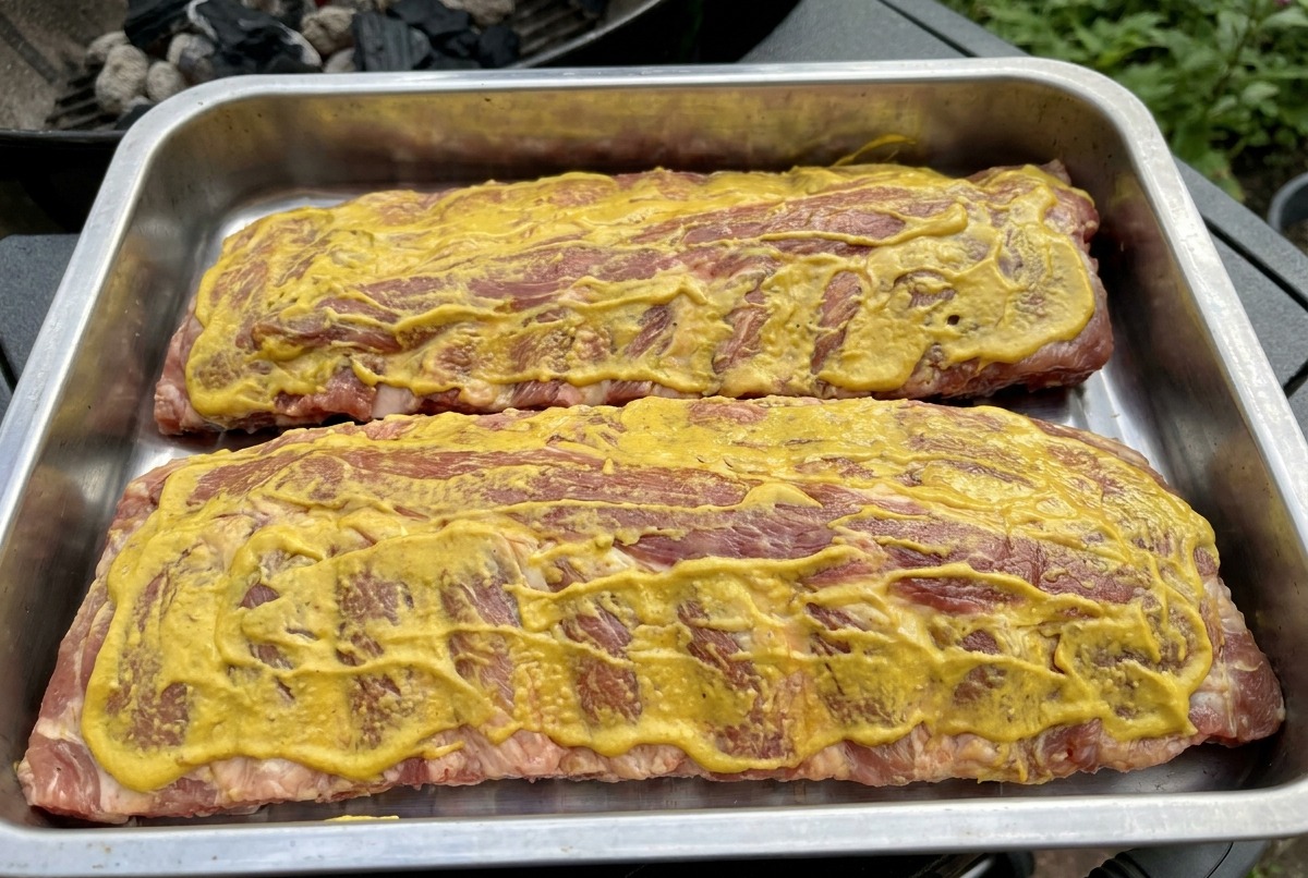Dos racks de costillas cubiertos con salsa amarilla sobre las rejillas de la parrilla, listos para ser ahumados.