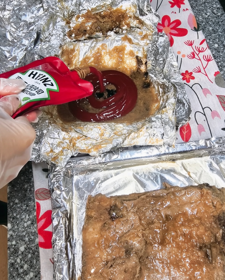 Kétchup siendo vertido en un cuenco que contiene jugos de cocción y otros ingredientes de la salsa.
