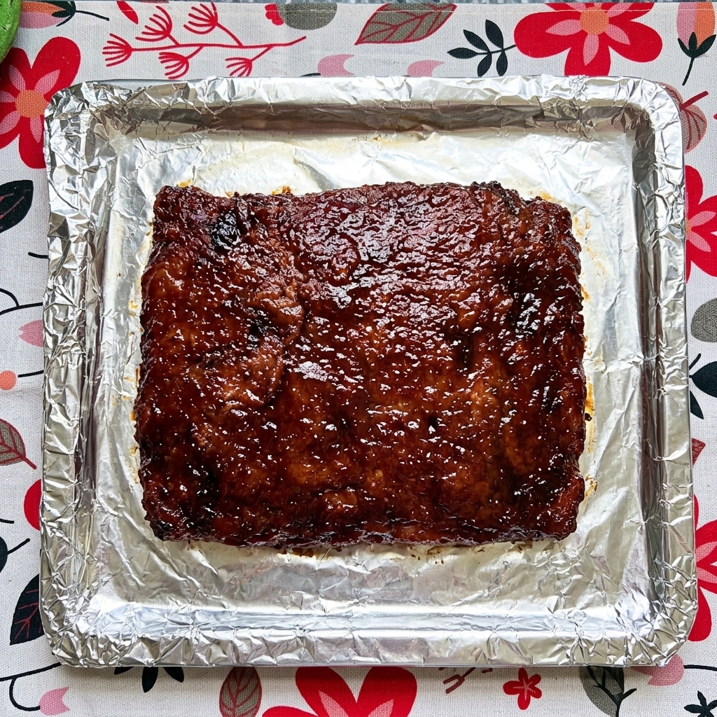Primer plano de un costillar de cerdo cocinado sobre papel aluminio siendo cubierto con una salsa oscura y brillante.