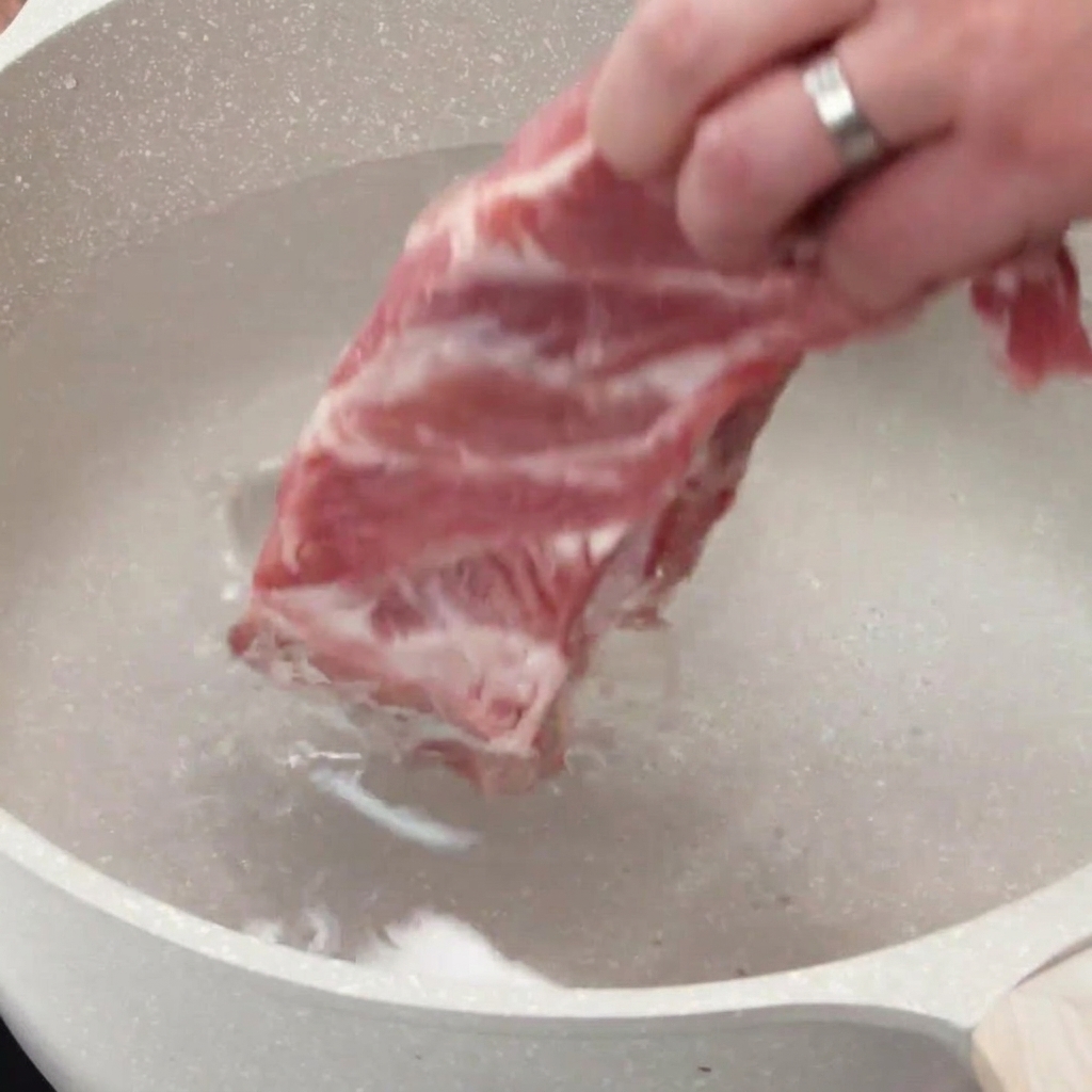 Una mano colocando un costillar de cerdo crudo en una olla llena de agua fría.