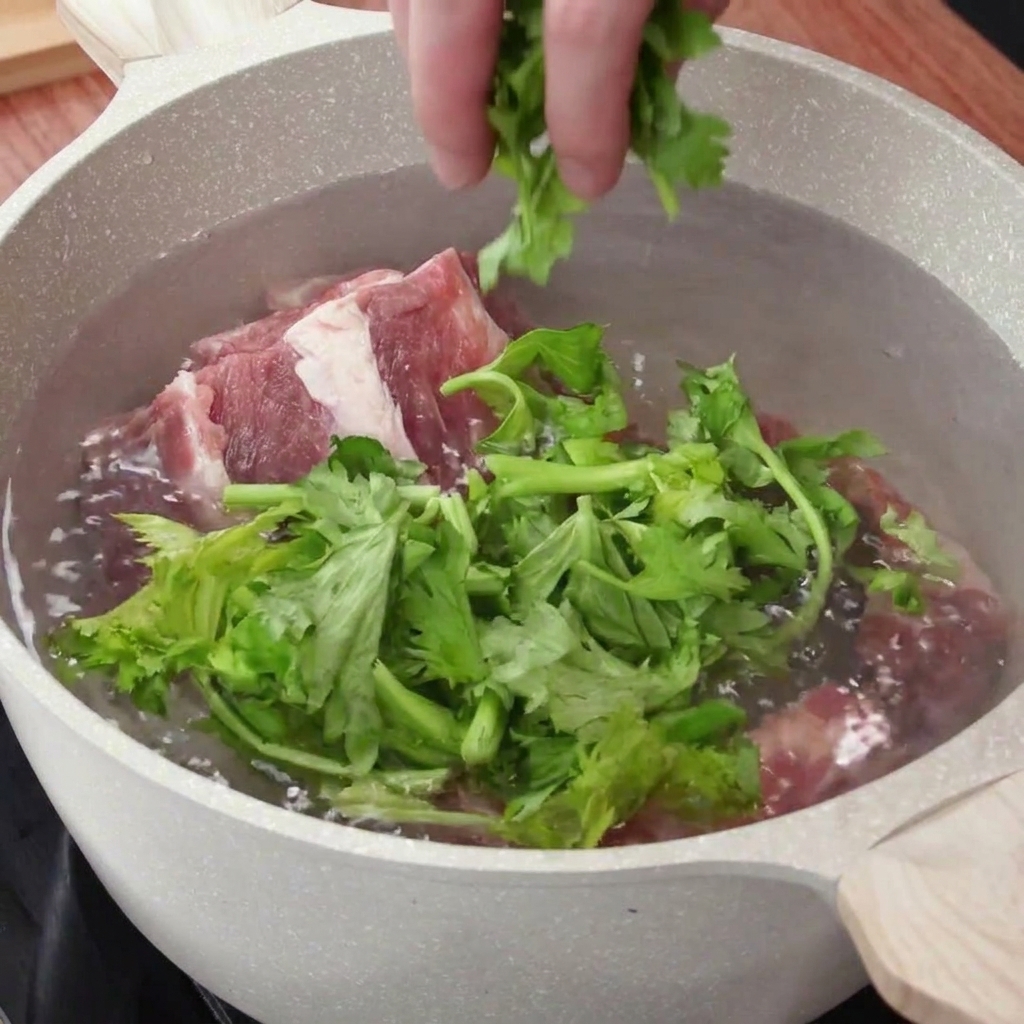 Una mano añadiendo hojas de apio y aromáticos picados en una olla de agua con costillas de cerdo crudas.