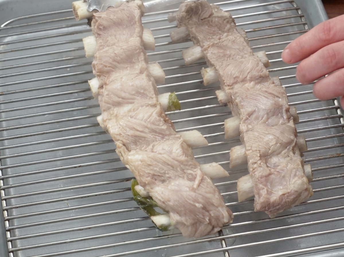 Costillares de cerdo cocidos descansando sobre una rejilla de metal con un plato colocado encima para aplanarlos.