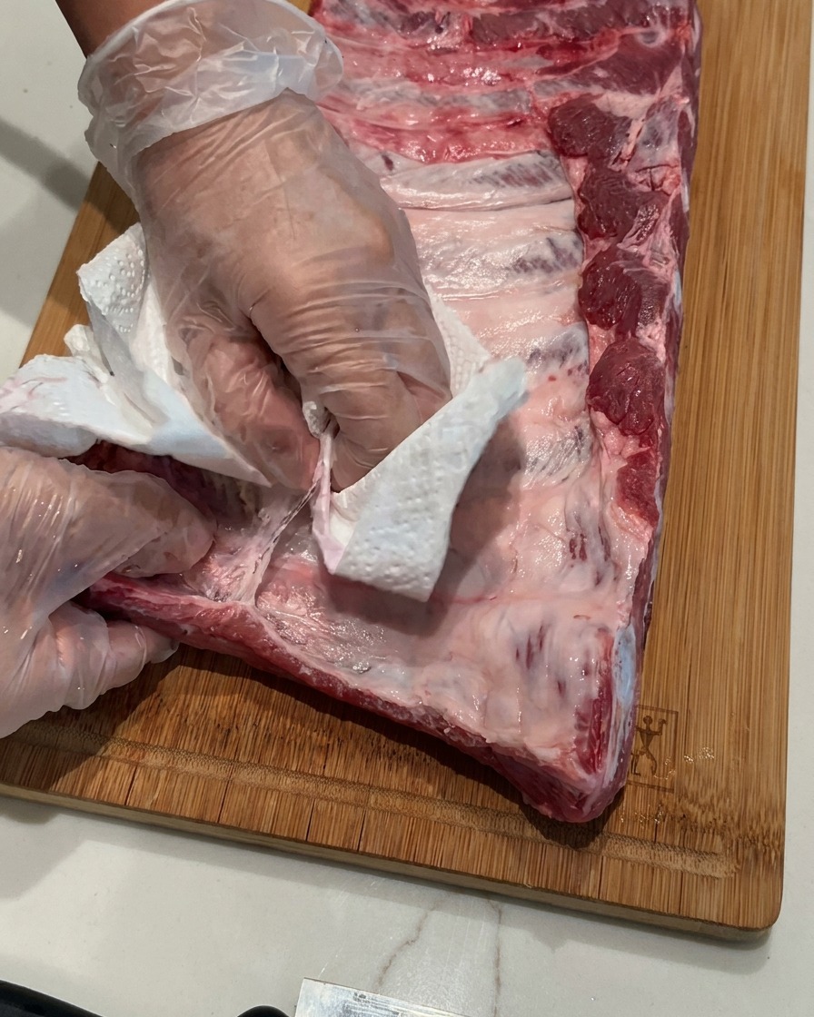 Manos con guantes de plástico transparentes usando una toalla de papel blanca para retirar una membrana delgada de un costillar de cerdo crudo en una tabla de cortar de madera.
