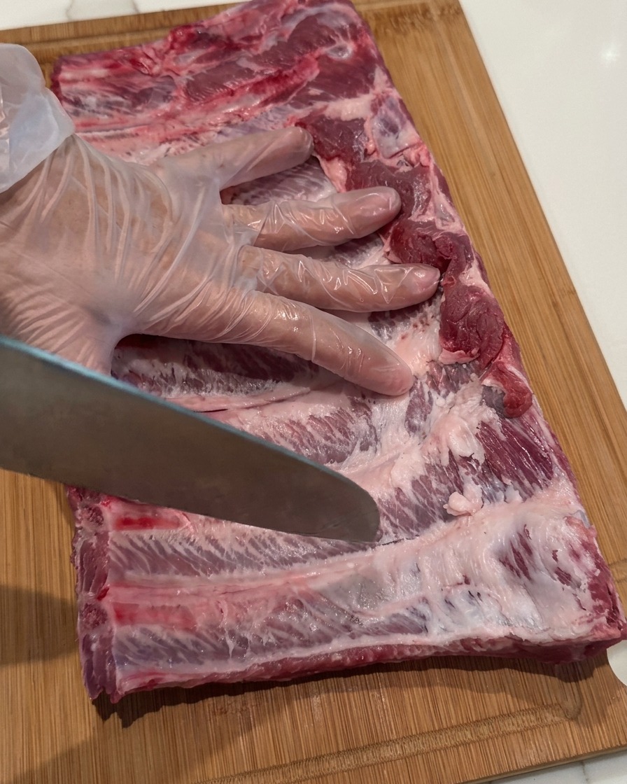 Una mano enguantada sosteniendo un cuchillo de chef, usando la punta para hacer pequeñas perforaciones en un costillar de cerdo crudo.