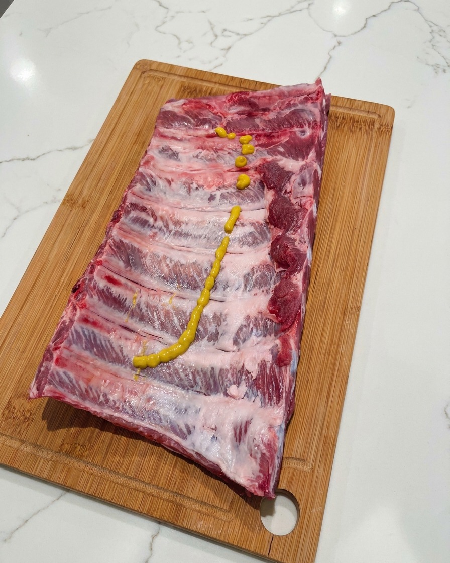 Una línea en zigzag de mostaza amarilla exprimida sobre la parte superior de un costillar de cerdo crudo sentado en una tabla de cortar de madera.