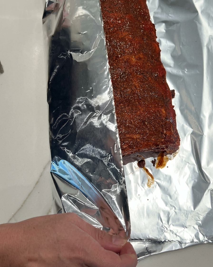 Una mano doblando cuidadosamente una pieza grande y brillante de papel de aluminio sobre un costillar de cerdo con salsa para sellarlas herméticamente.
