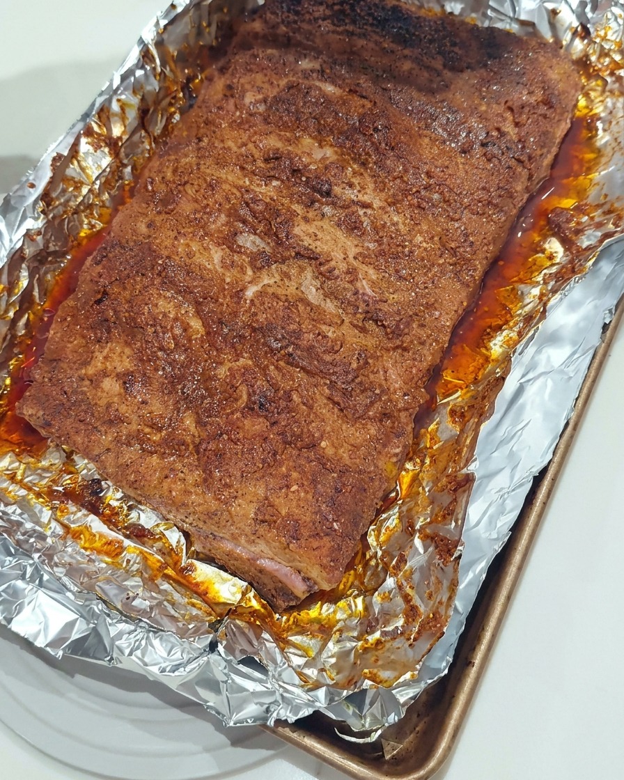 Un cepillo de silicona aplicando una capa brillante de salsa BBQ espesa sobre la superficie de costillas de cerdo asadas en una bandeja forrada con papel de aluminio.