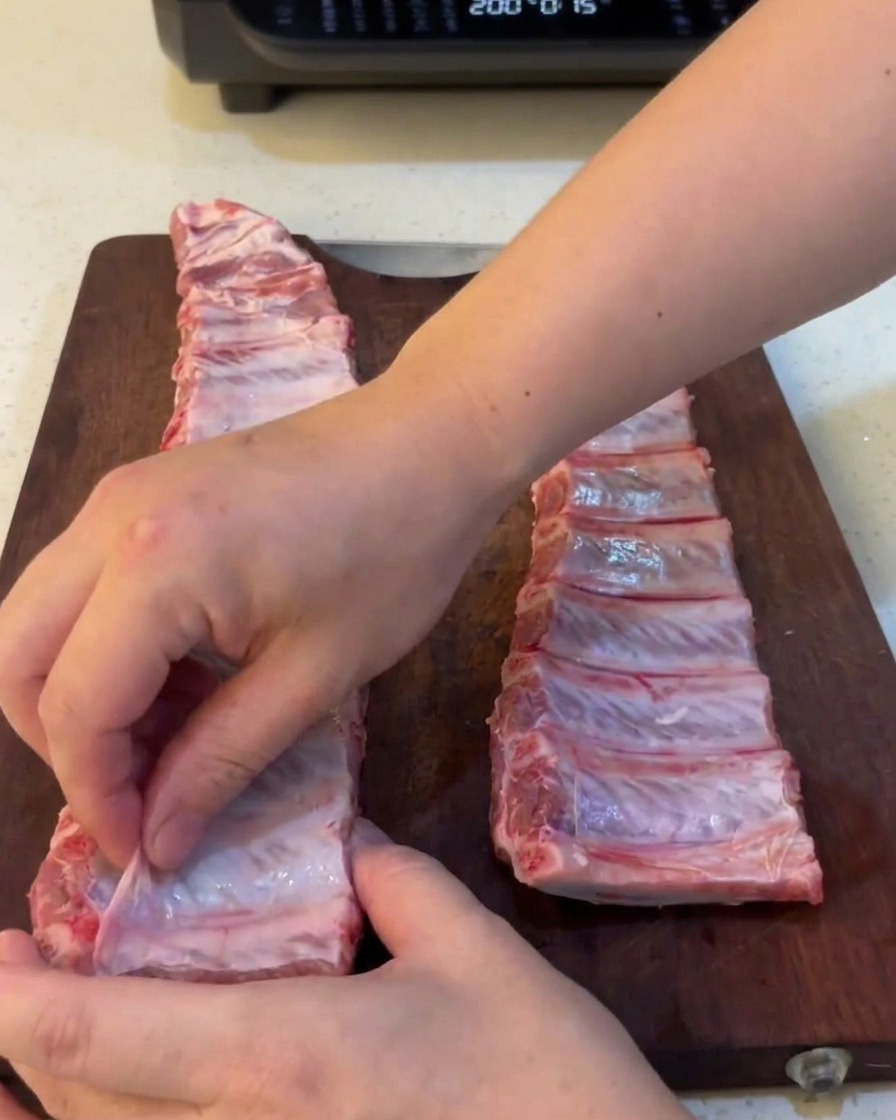 Primer plano de unas manos retirando una membrana fina y traslúcida de la parte posterior de costillas de cerdo crudas.