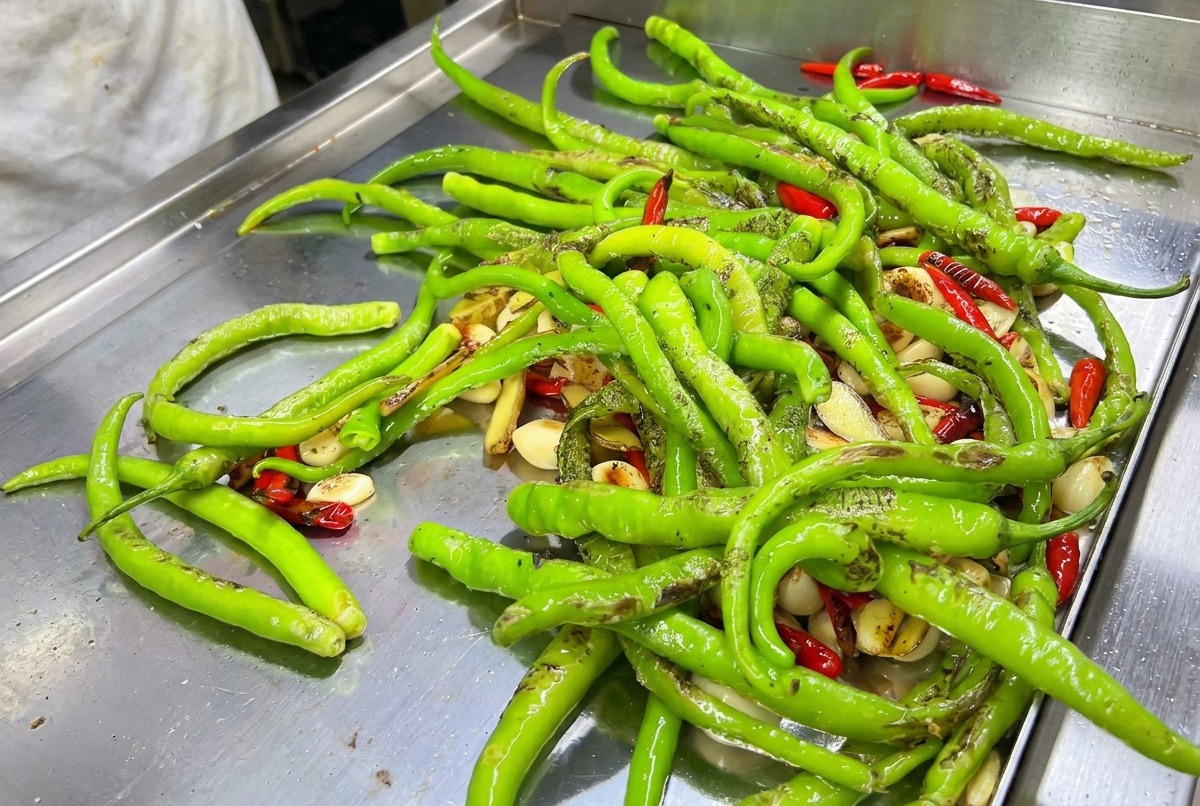 Una mezcla de chiles verdes y rojos siendo quemados con soplete sobre una plancha caliente.