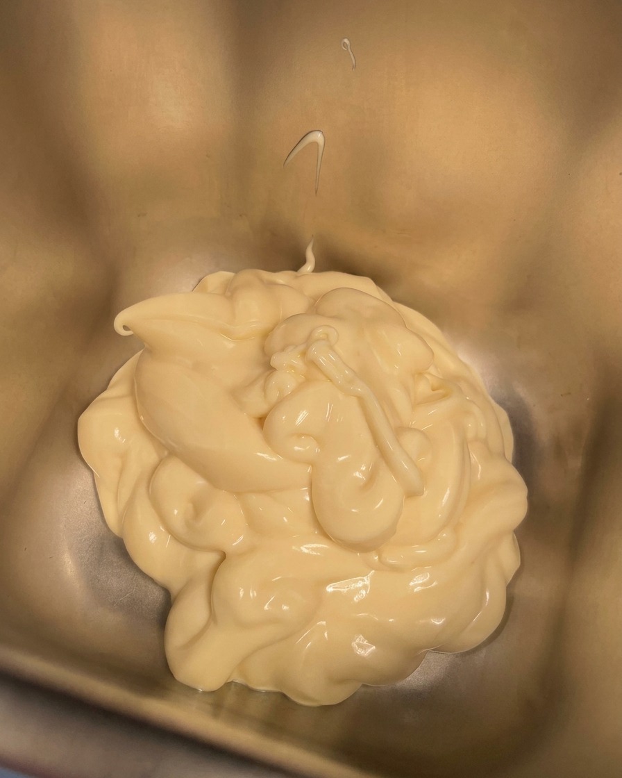 Un tazón pequeño con crema siendo vertida en un recipiente metálico que ya contiene mayonesa cremosa.