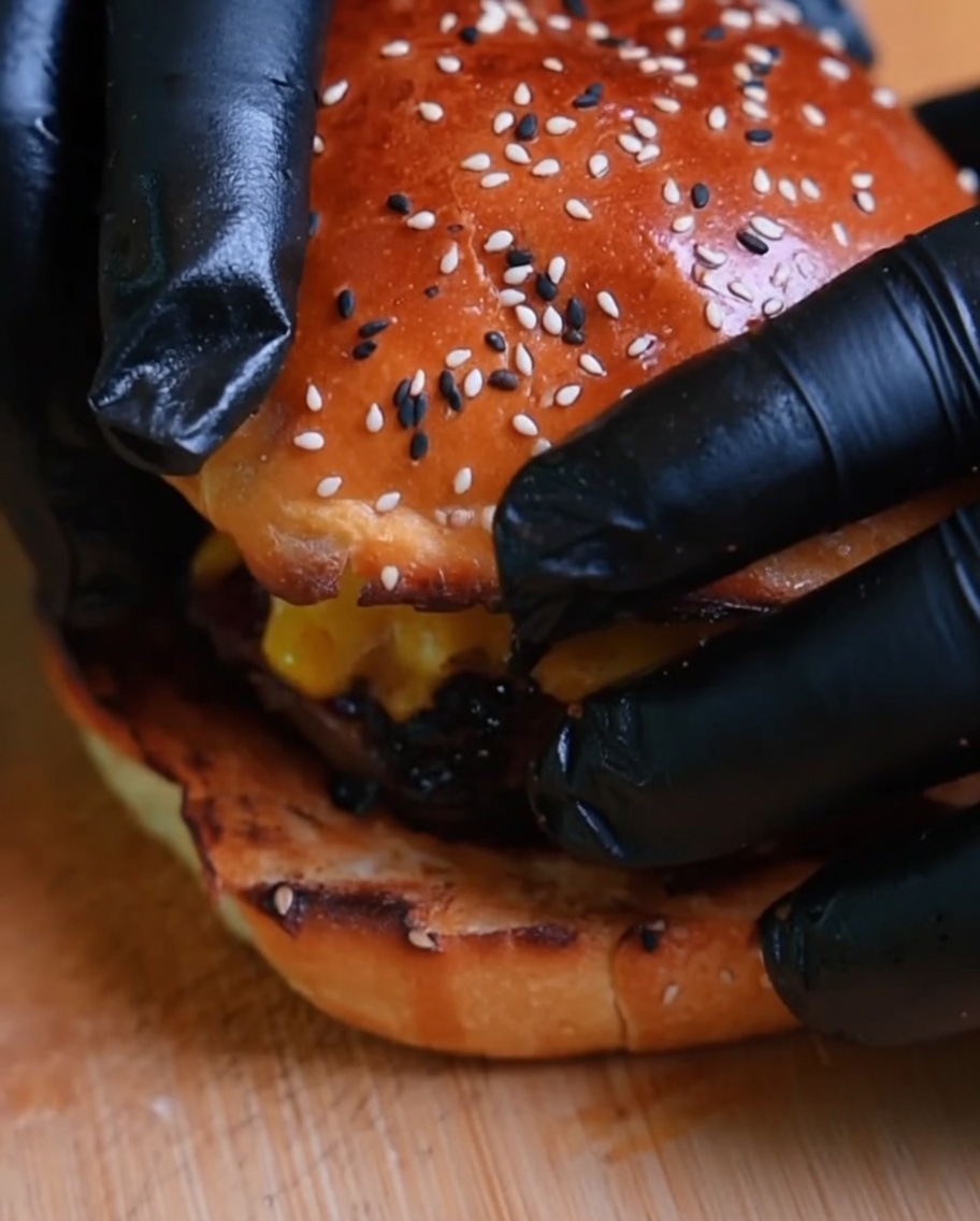 Primer plano de una hamburguesa siendo finalizada al colocar el pan superior, sostenida por manos con guantes.