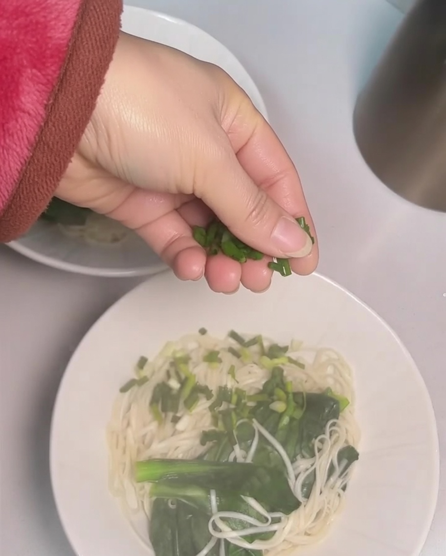Mano espolvoreando cebolletas picadas sobre un tazón de fideos.