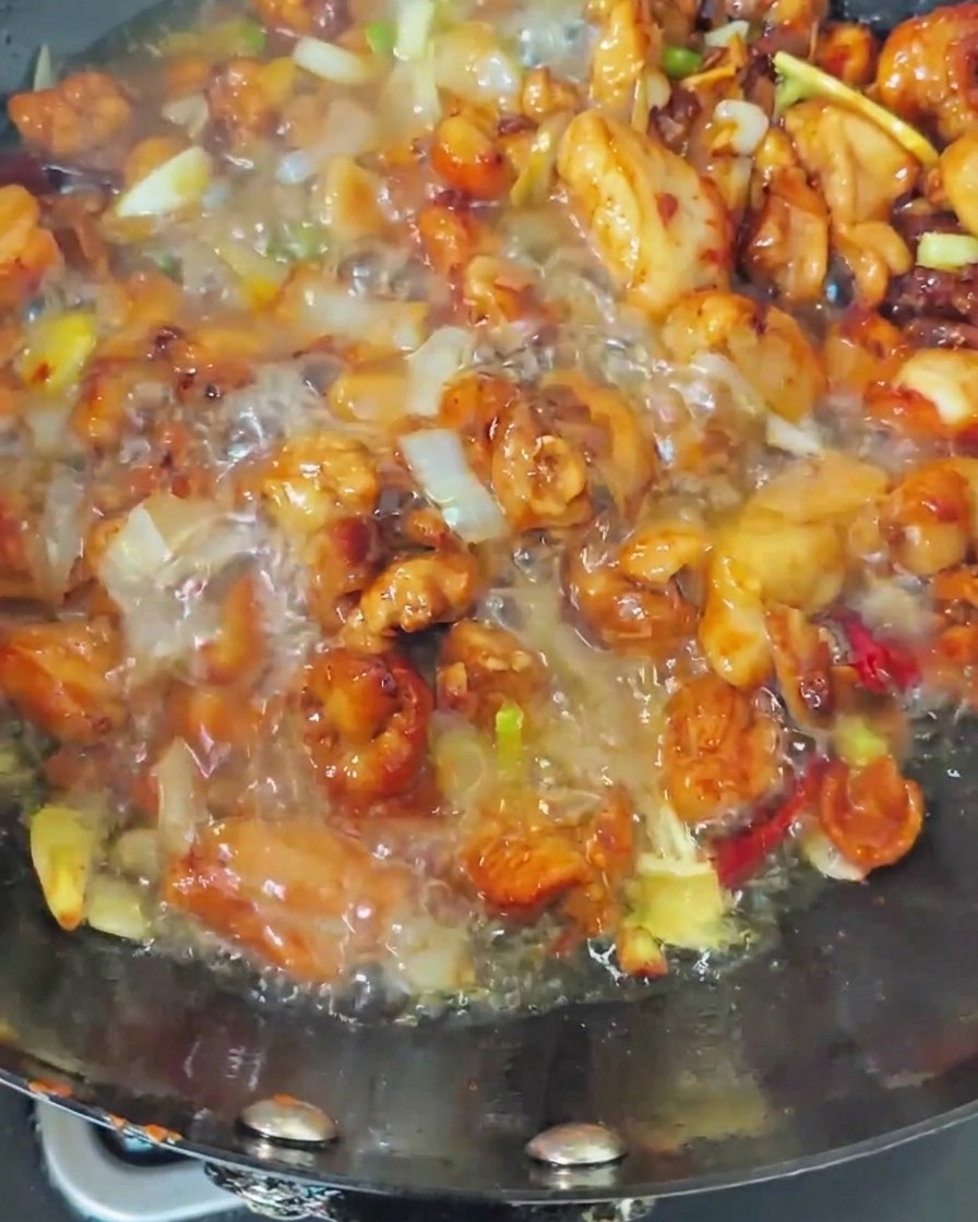 Agua hirviendo siendo vertida en un wok que contiene pollo sellado y aromáticos, creando un caldo burbujeante.