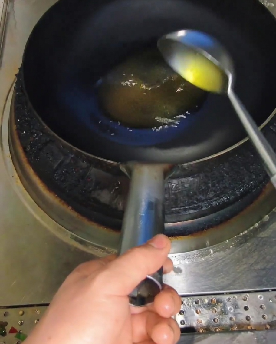 Un cucharón metálico vertiendo aceite amarillo en un wok oscuro y caliente.