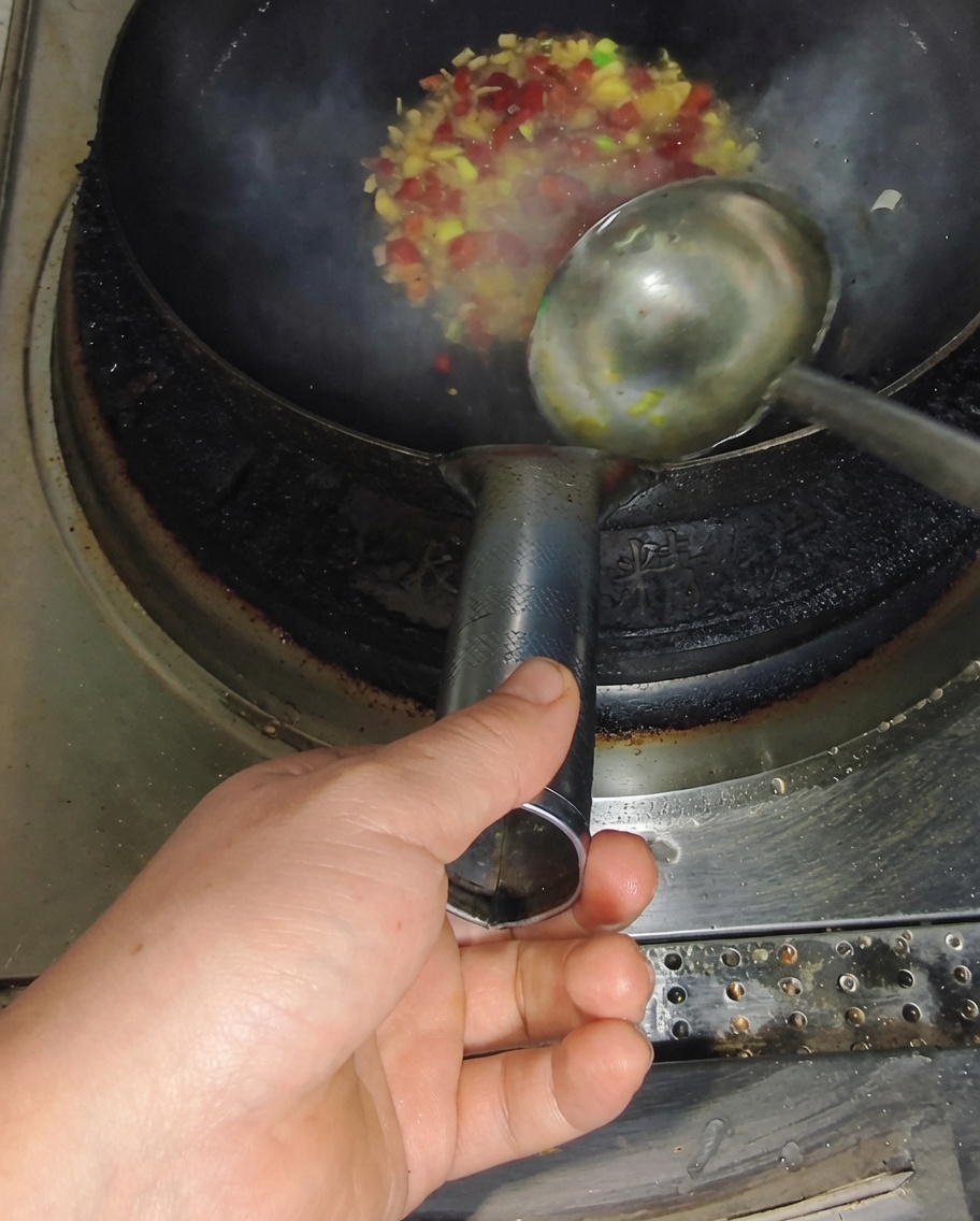 Un cucharón vertiendo caldo claro en un wok caliente con chiles sofritos y cebollino, creando vapor.