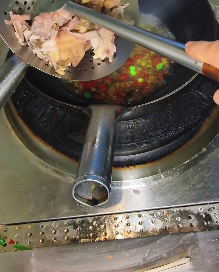 Una cuchara ranurada añadiendo trozos de pollo cocido desmenuzado en un wok con caldo picante burbujeante.