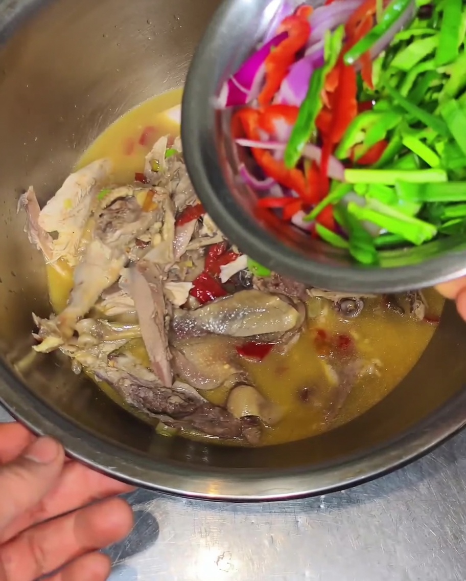 Un bol pequeño metálico vertiendo cebolla roja, pimientos verdes y rojos sobre pollo y caldo en un bol grande.