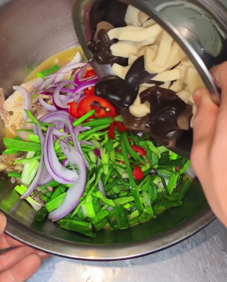 Manos inclinando un bol pequeño para añadir hongos oreja de madera y tiras de piel de tofu en un bol grande con pollo, pimientos y cebollas.