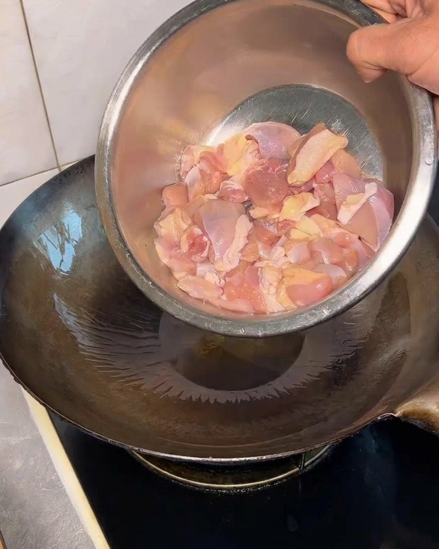 Trozos de contramuslo de pollo crudo siendo vertidos desde un bol de metal a un wok caliente con aceite.