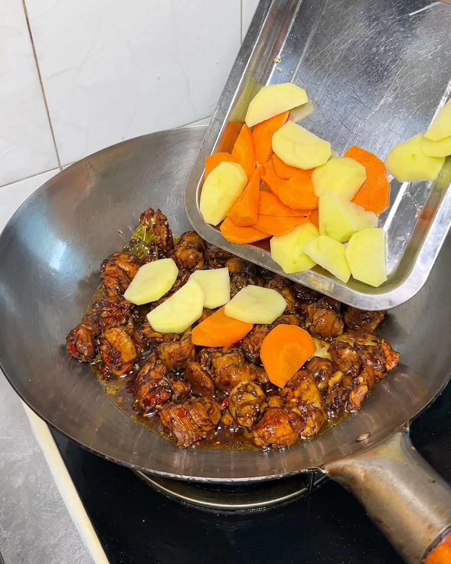 Trozos de patata y zanahoria siendo vertidos en un wok con pollo en salsa oscura.