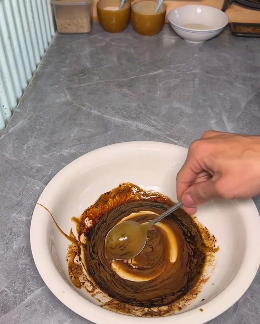 Una mano usando una cuchara para mezclar una salsa oscura y rica de pasta de sésamo y soja en un bol blanco.