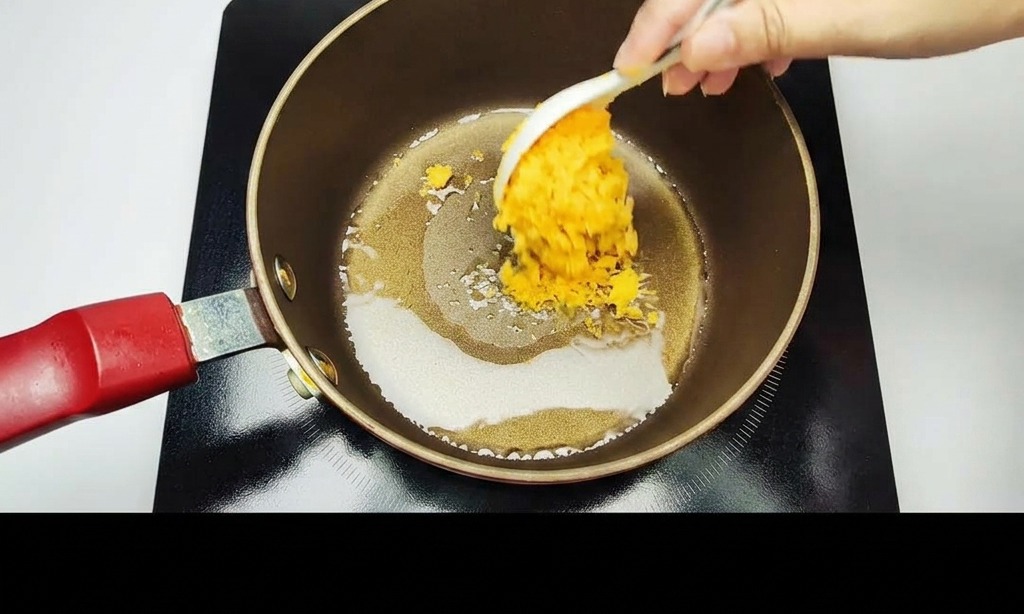 Una mano usando una cuchara blanca para sofreír yemas de huevo saladas doradas machacadas en una olla antiadherente oscura.