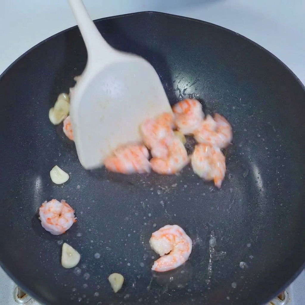 Piezas de okra siendo salteadas en una sartén con gambas y ajo usando una espátula blanca.