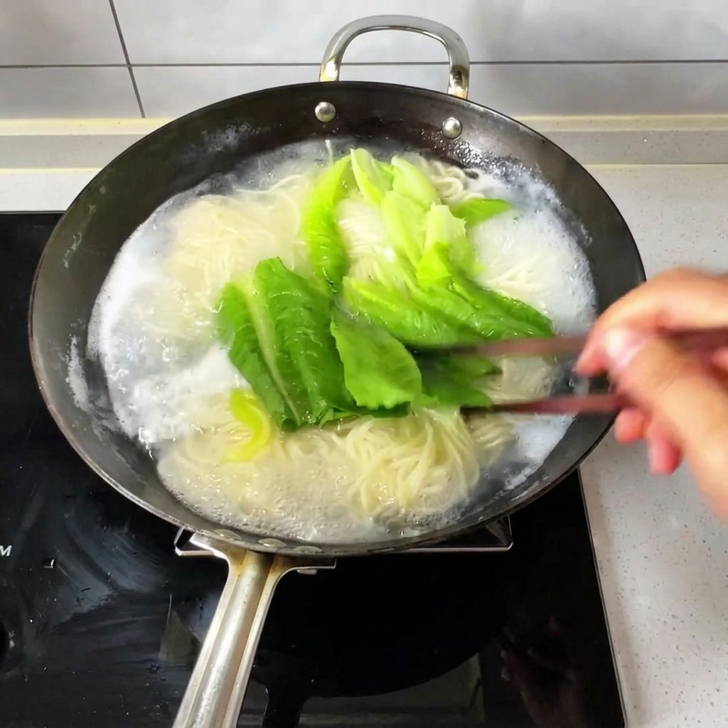 Fideos finos y verduras de hoja verde hirviendo en un wok de agua burbujeante, removidos con palillos de madera.