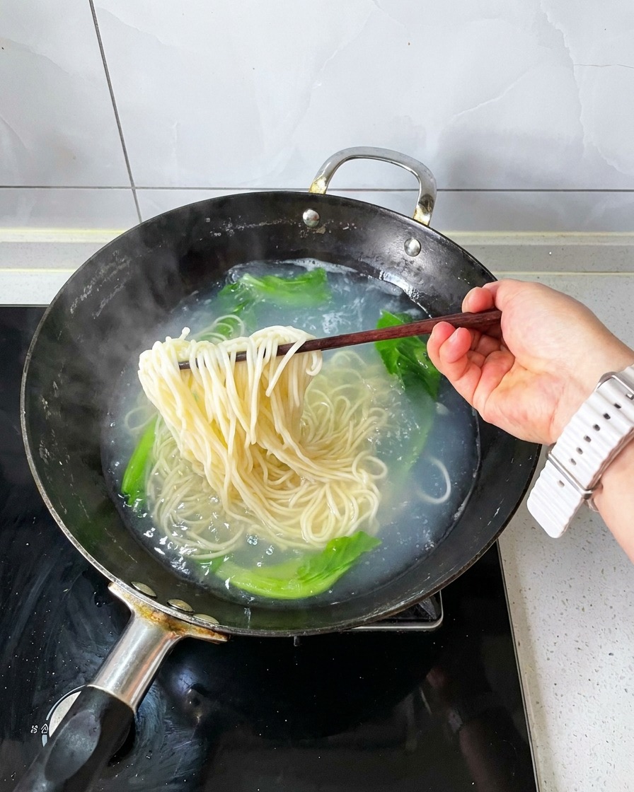 Palillos de madera levantando un manojo de fideos cocidos y verduras blanqueadas fuera del agua hirviendo.