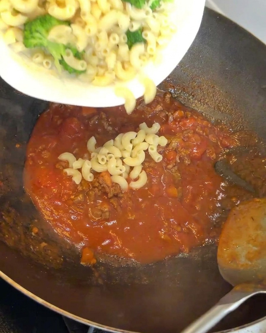 Macarrones y brócoli siendo añadidos a un wok lleno de una rica salsa de carne.